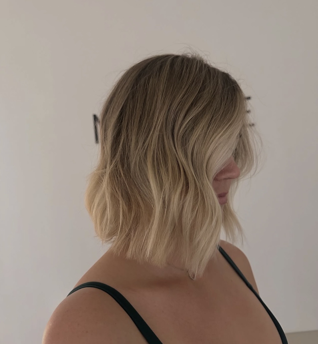 Side view of a woman with shoulder-length wavy blonde hair, wearing a black strap top, against a plain background.
