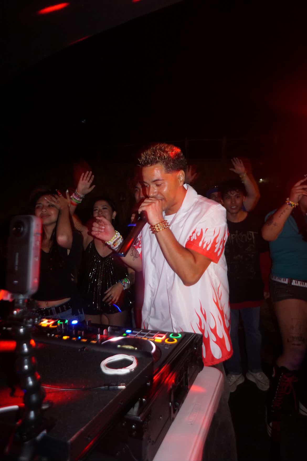 Person with curly hair singing into a microphone at a DJ booth, surrounded by people dancing in a dark club.
