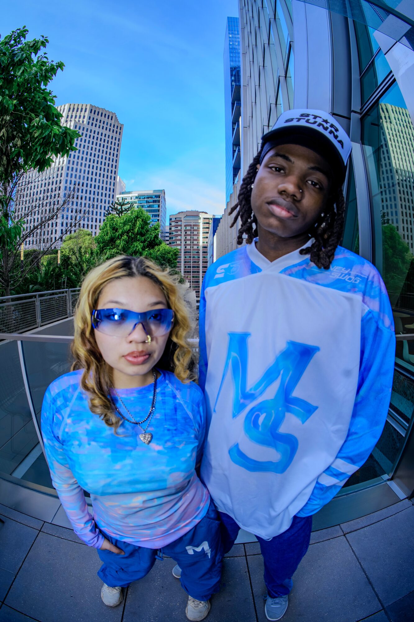 Two young people stand outdoors in an urban setting with tall buildings and trees, one wearing sunglasses and a cap.