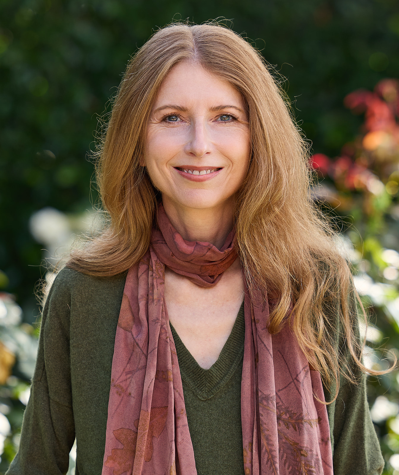 Woman with long, wavy hair smiling outdoors, wearing a green sweater and pink scarf, with blurred greenery background.