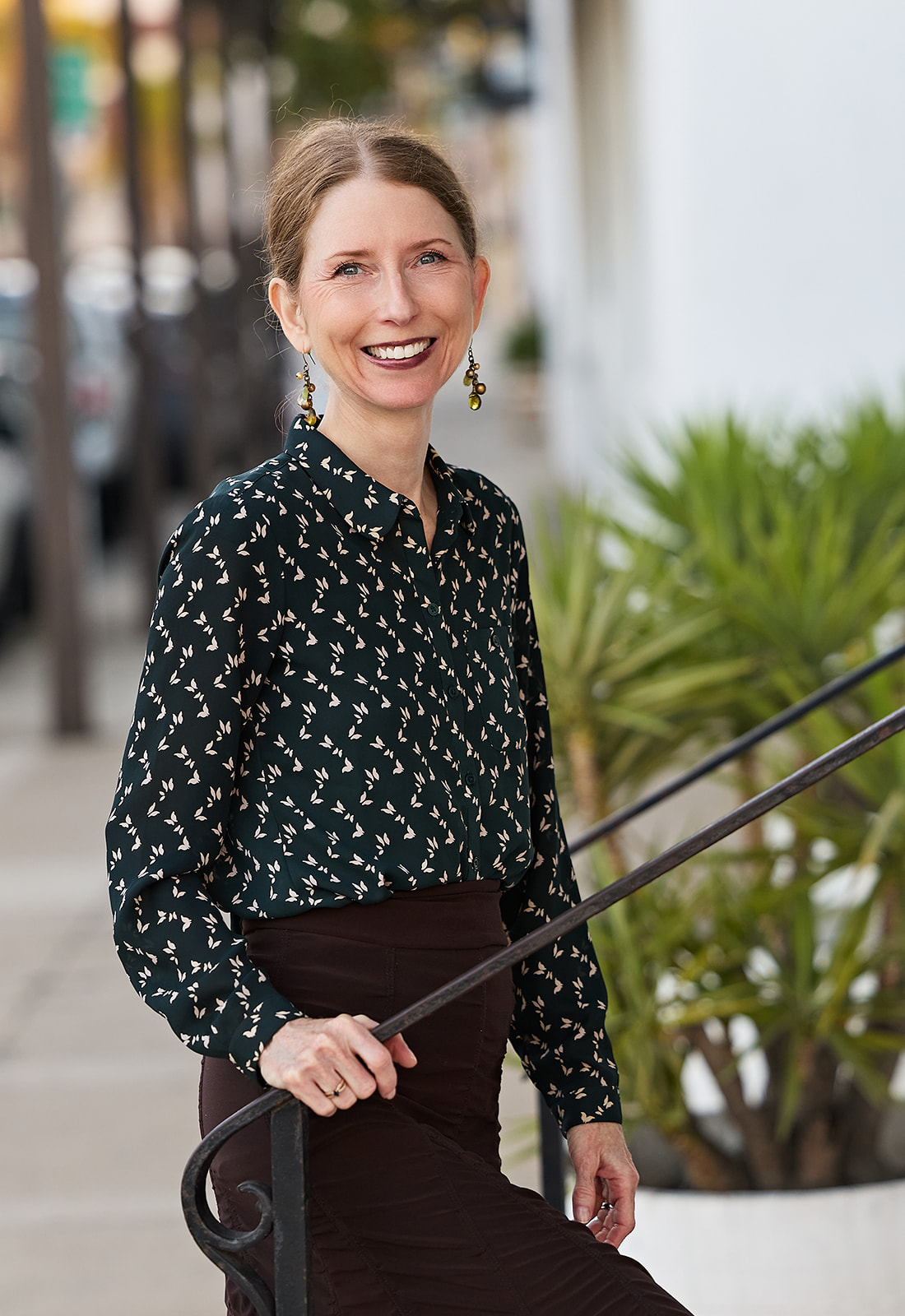 Smiling woman with earrings, wearing a patterned blouse and dark skirt, standing outdoors near plants and railing.