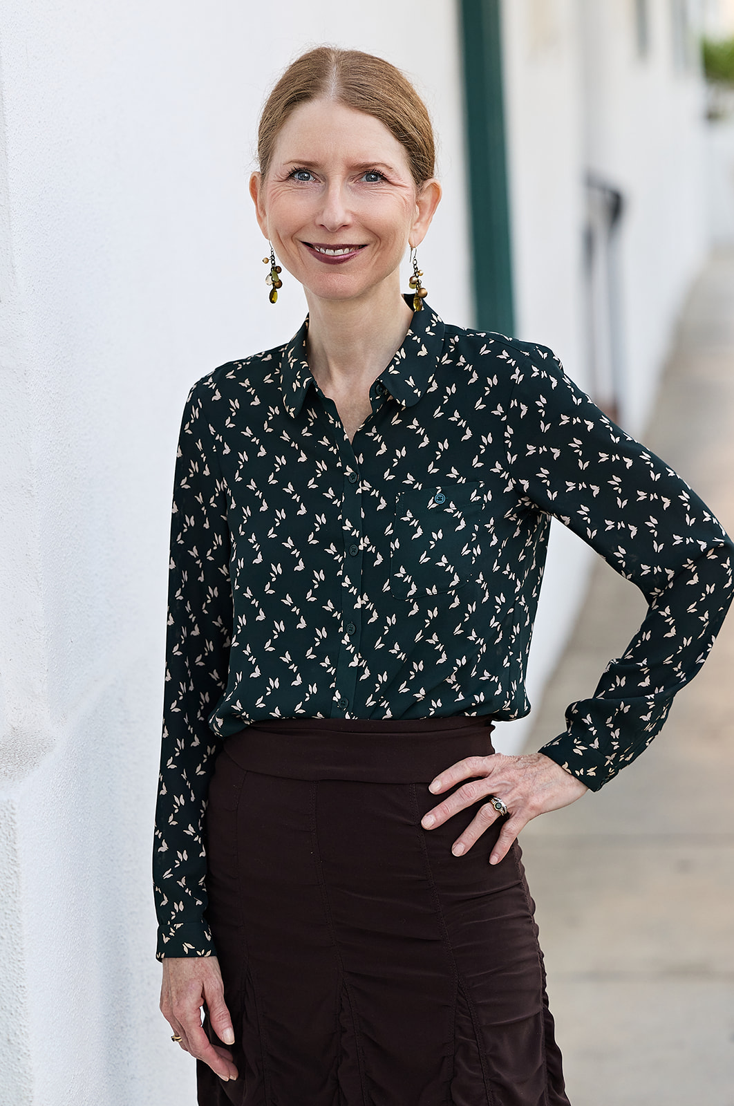 Woman with light brown hair in a bun, wearing a black patterned blouse and dark skirt, standing outdoors smiling.
