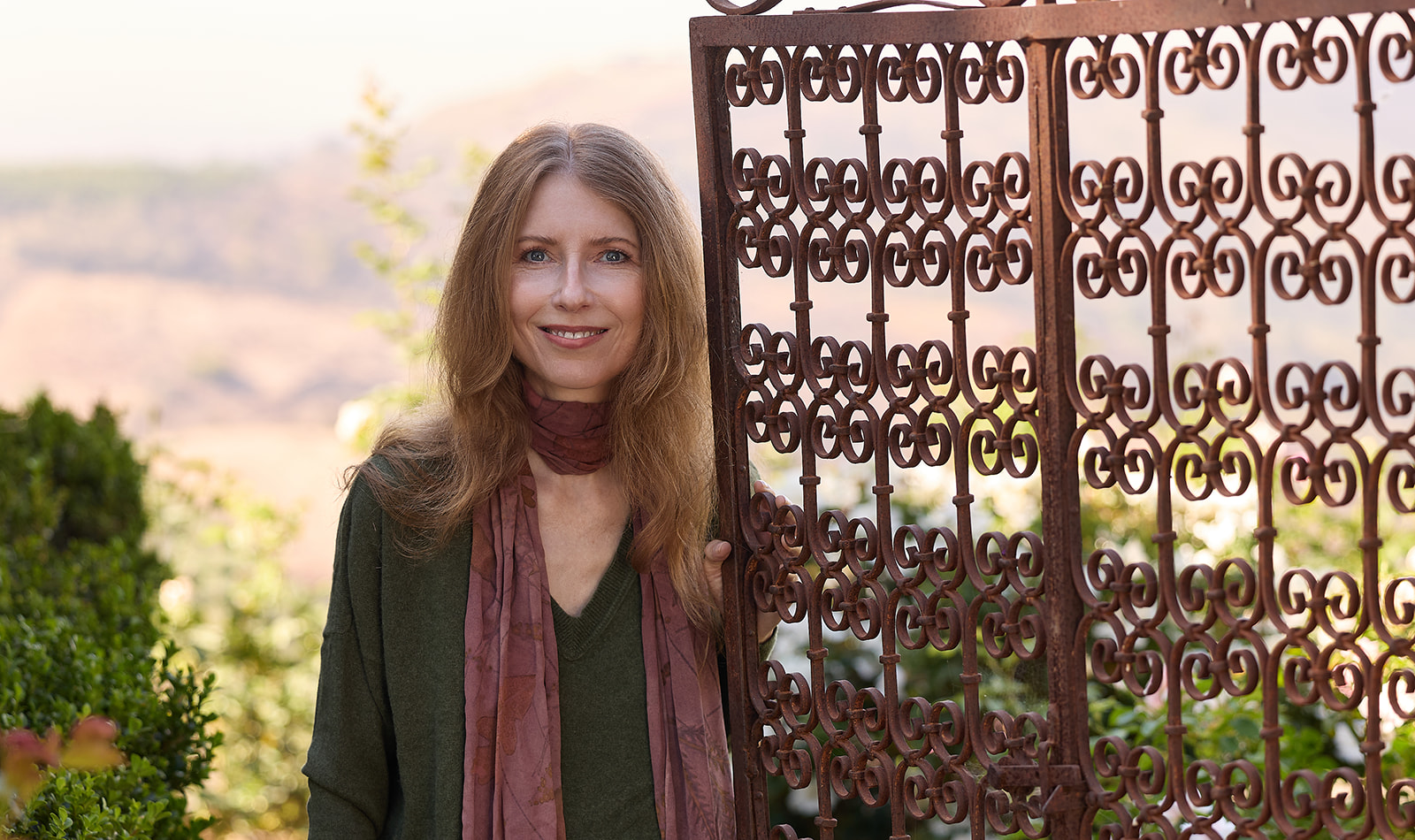 Woman with long hair smiling outdoors next to a decorative metal gate, greenery, and a landscape background.