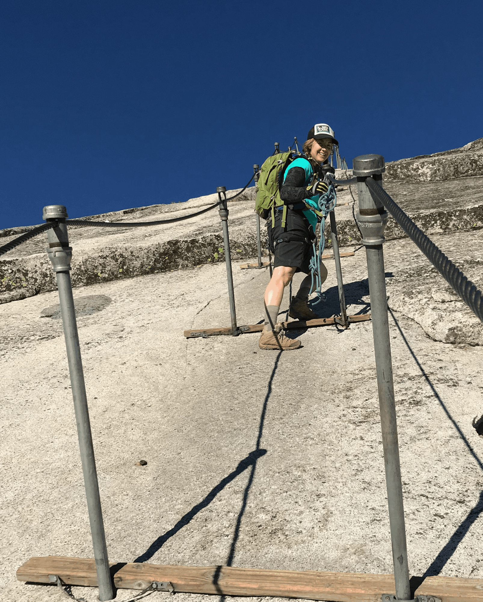 Person with a backpack and hat climbing a steep outdoor trail with metal railings, against a clear blue sky.
