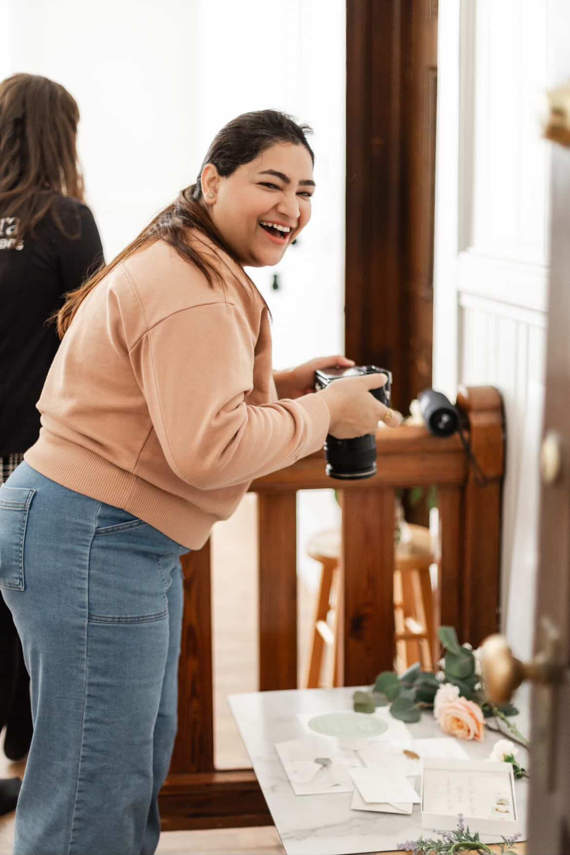 Young woman laughing, holding a camera, standing near a table with flowers and papers, in a bright room.