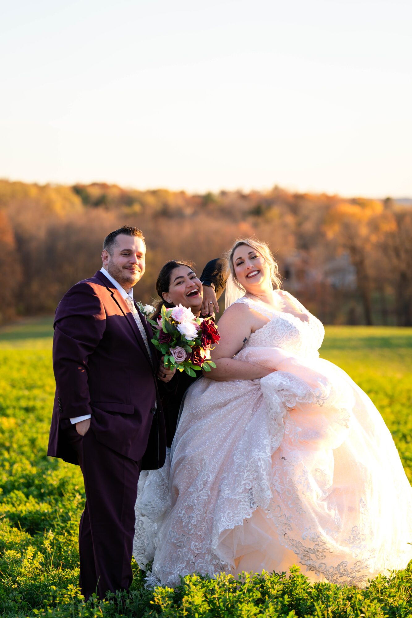 Three people outdoors in a field, one in a wedding dress, holding a bouquet, smiling, with a landscape background.
