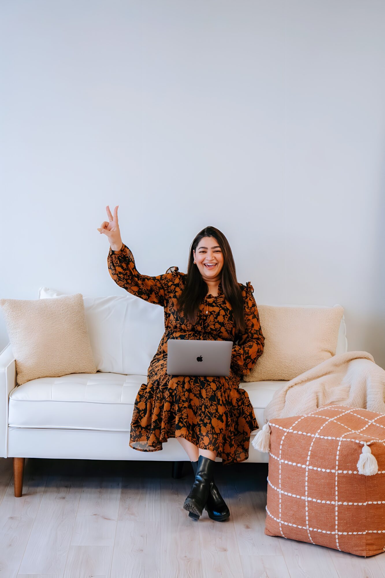 Woman sitting on a white sofa, smiling, holding a laptop, raising two fingers in a peace sign, in a bright room.