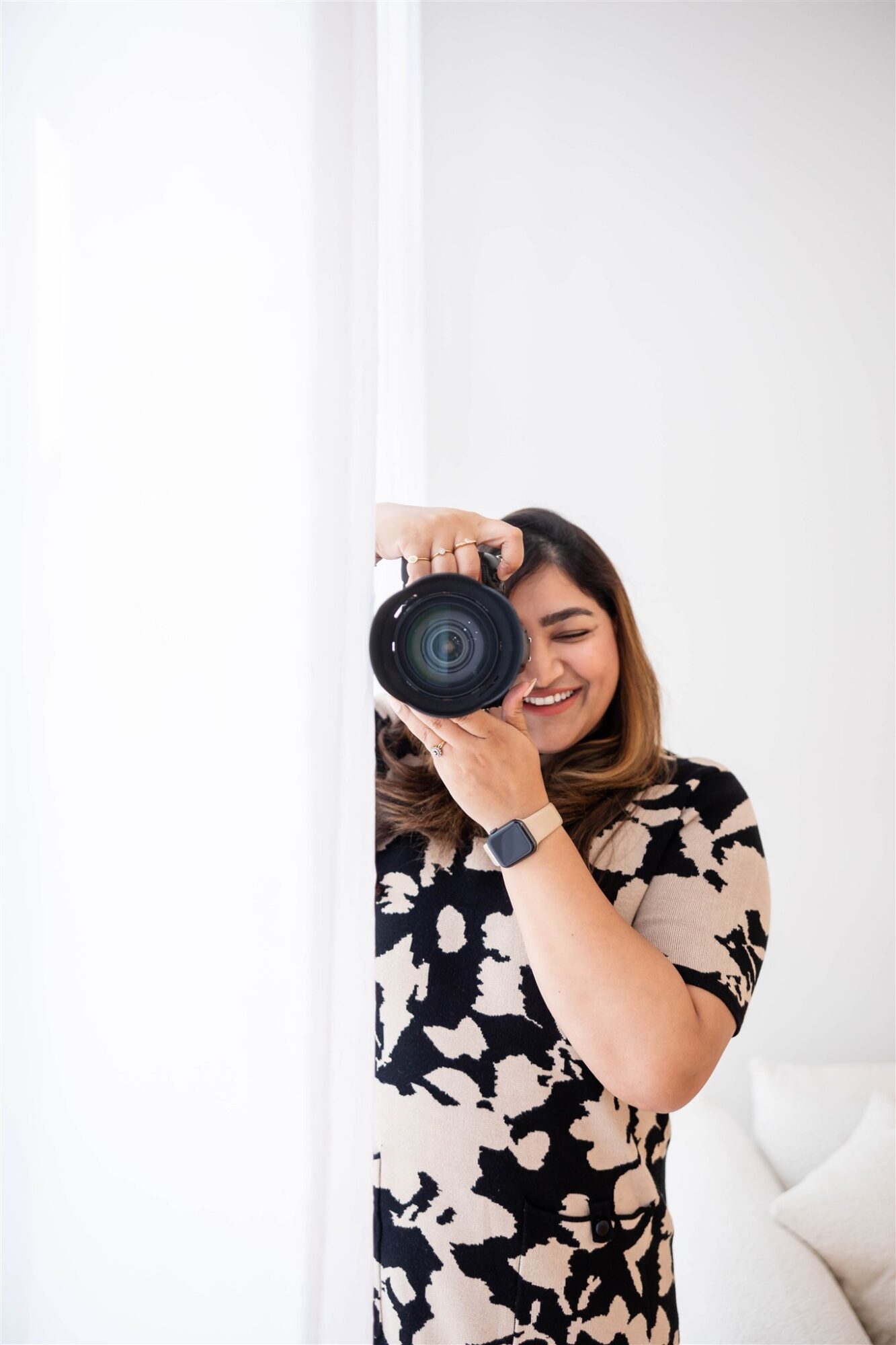 Woman taking a selfie with a camera, smiling, in a bright room with white walls and a pillow visible.