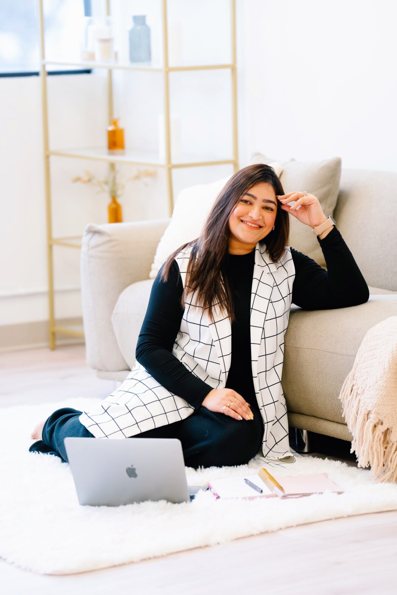 Young woman sitting on a rug with a laptop, smiling, in a bright room with a sofa and shelves in the background.