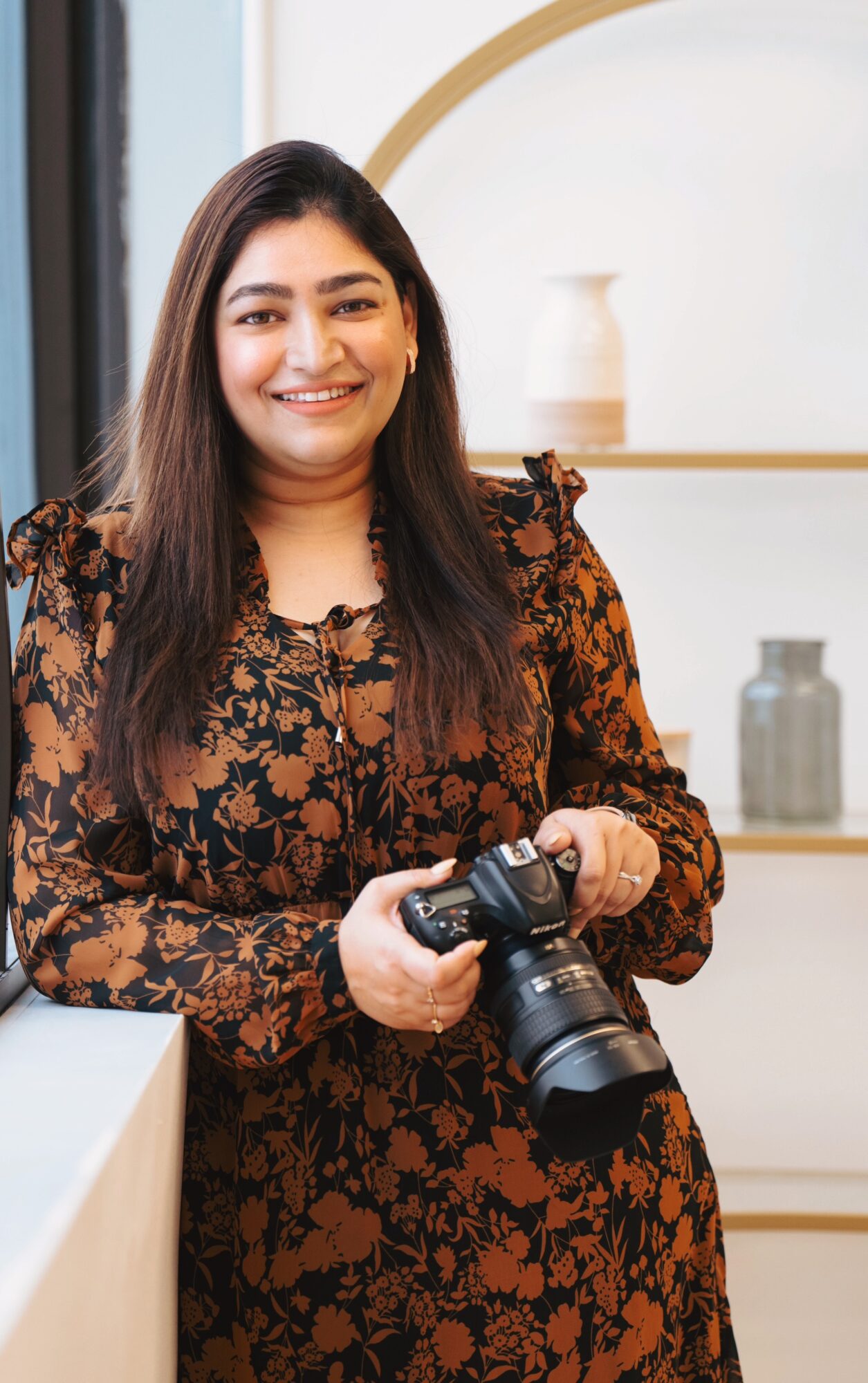 Smiling woman holding a camera indoors, wearing a floral dress, with shelves and decorative items in the background.