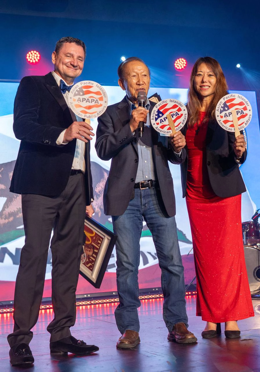 Three people on stage holding circular signs, smiling, with colorful lights and a screen in the background.