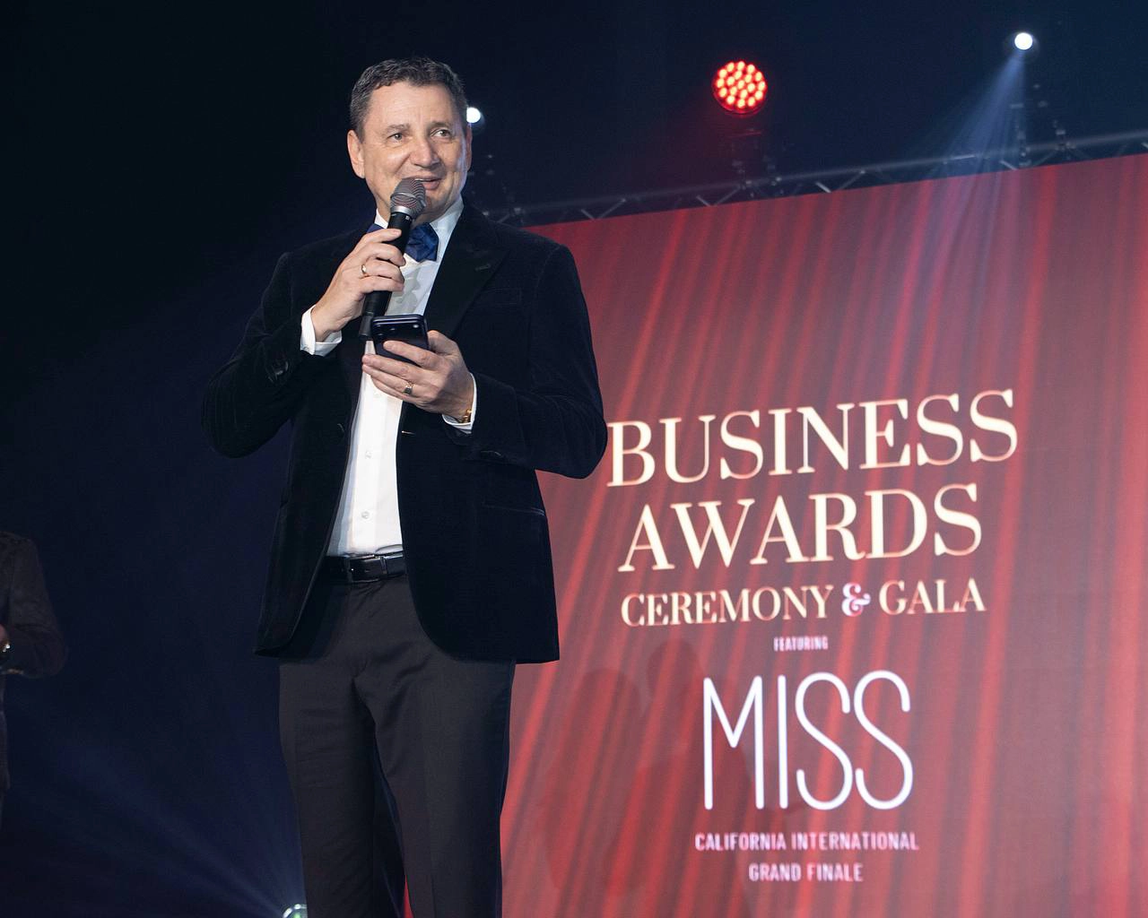 Man in tuxedo speaking into microphone on stage at business awards event, red curtain background.