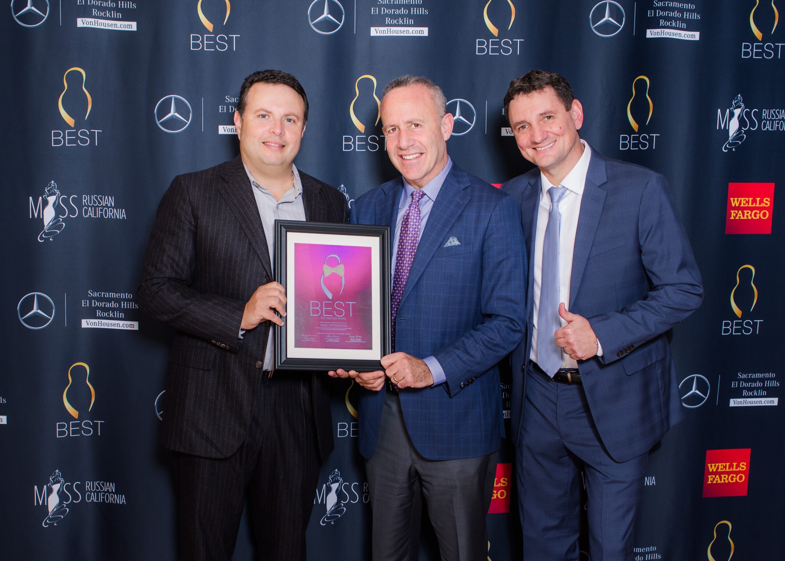 Three men in suits holding an award plaque at an event with a blue backdrop featuring logos.