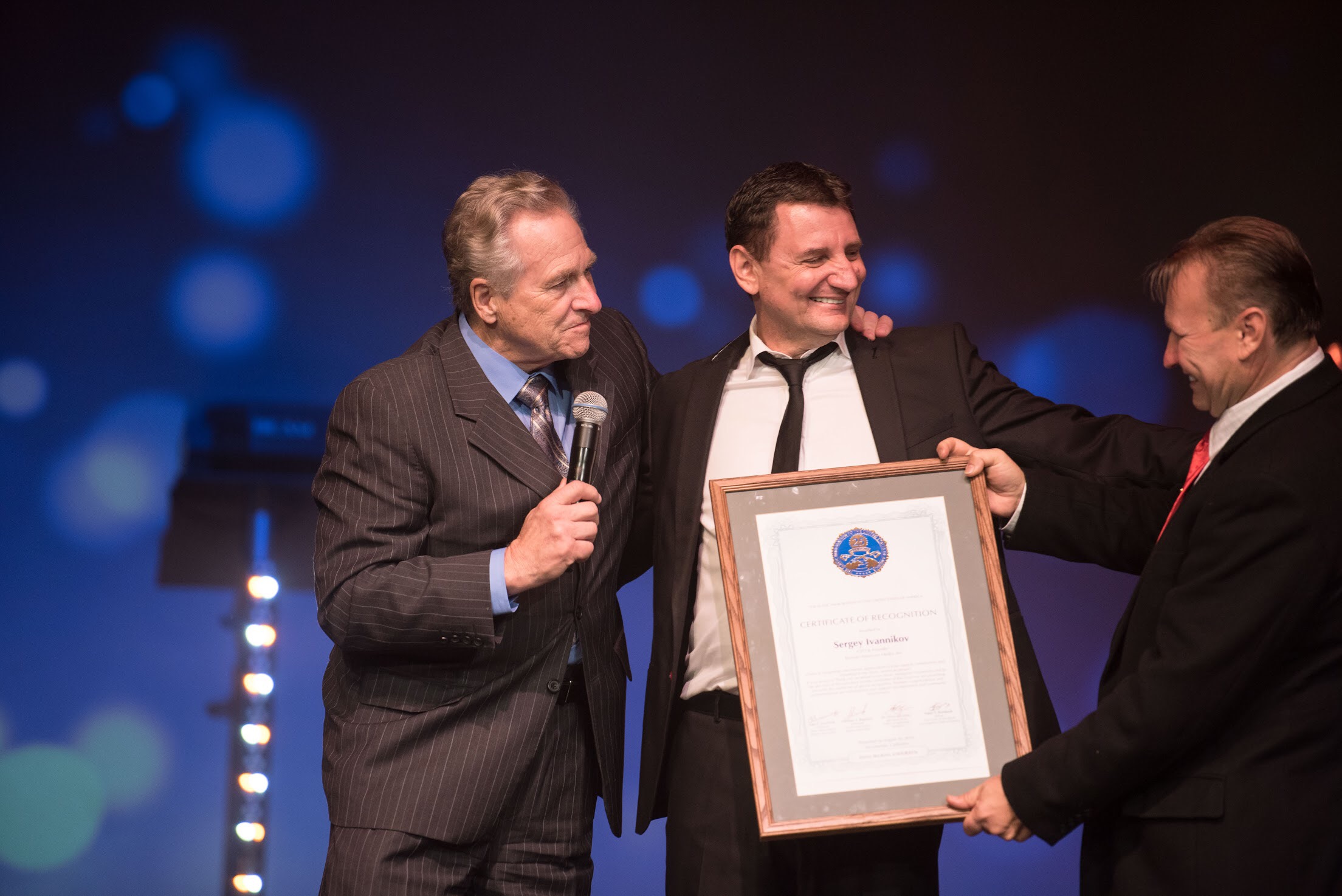 Three men in suits on stage, one holding a framed certificate, with a dark background and blue lights, smiling.