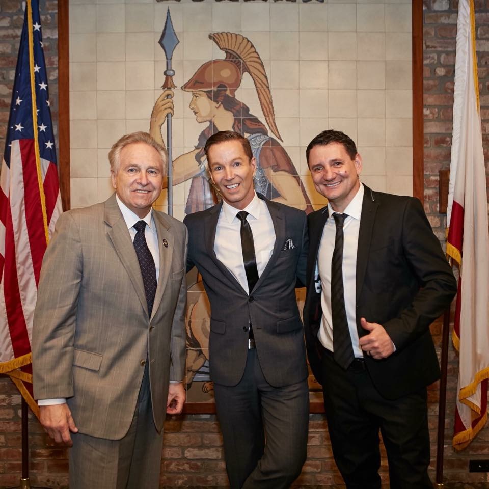 Three men in suits standing in front of a mural of a warrior with a spear, flags on either side, smiling.