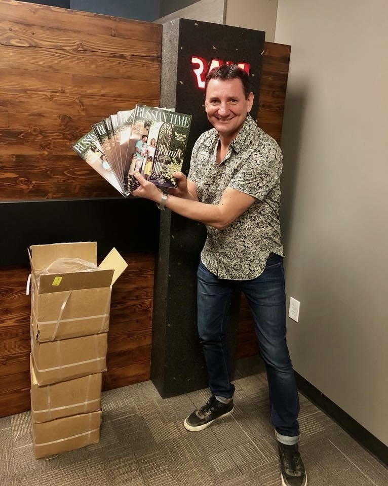 Person smiling holding multiple magazines, standing next to a cardboard box, in an indoor setting with wooden and gray walls.