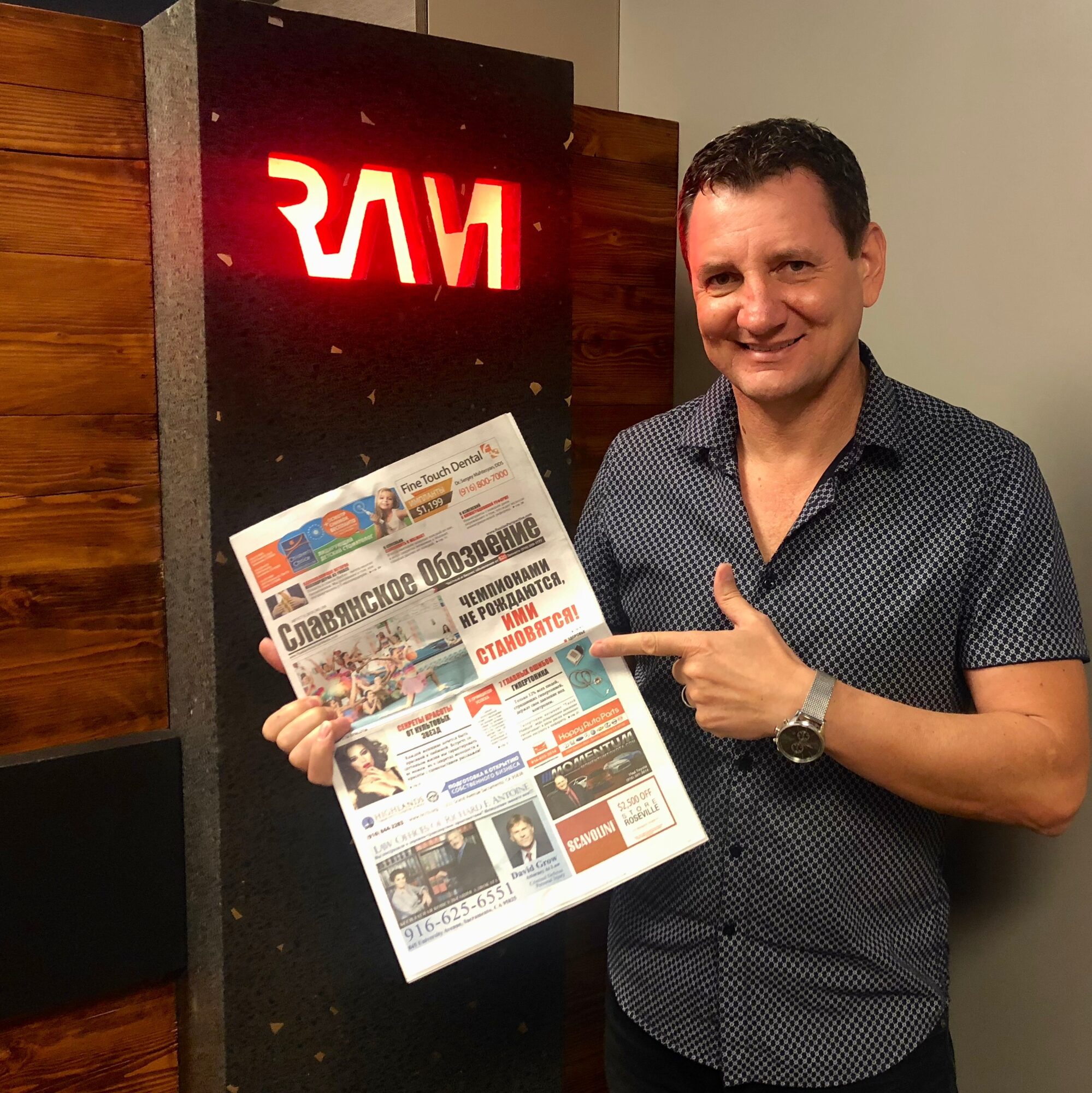 Man smiling and pointing at a newspaper, standing next to a RAM sign, indoors.