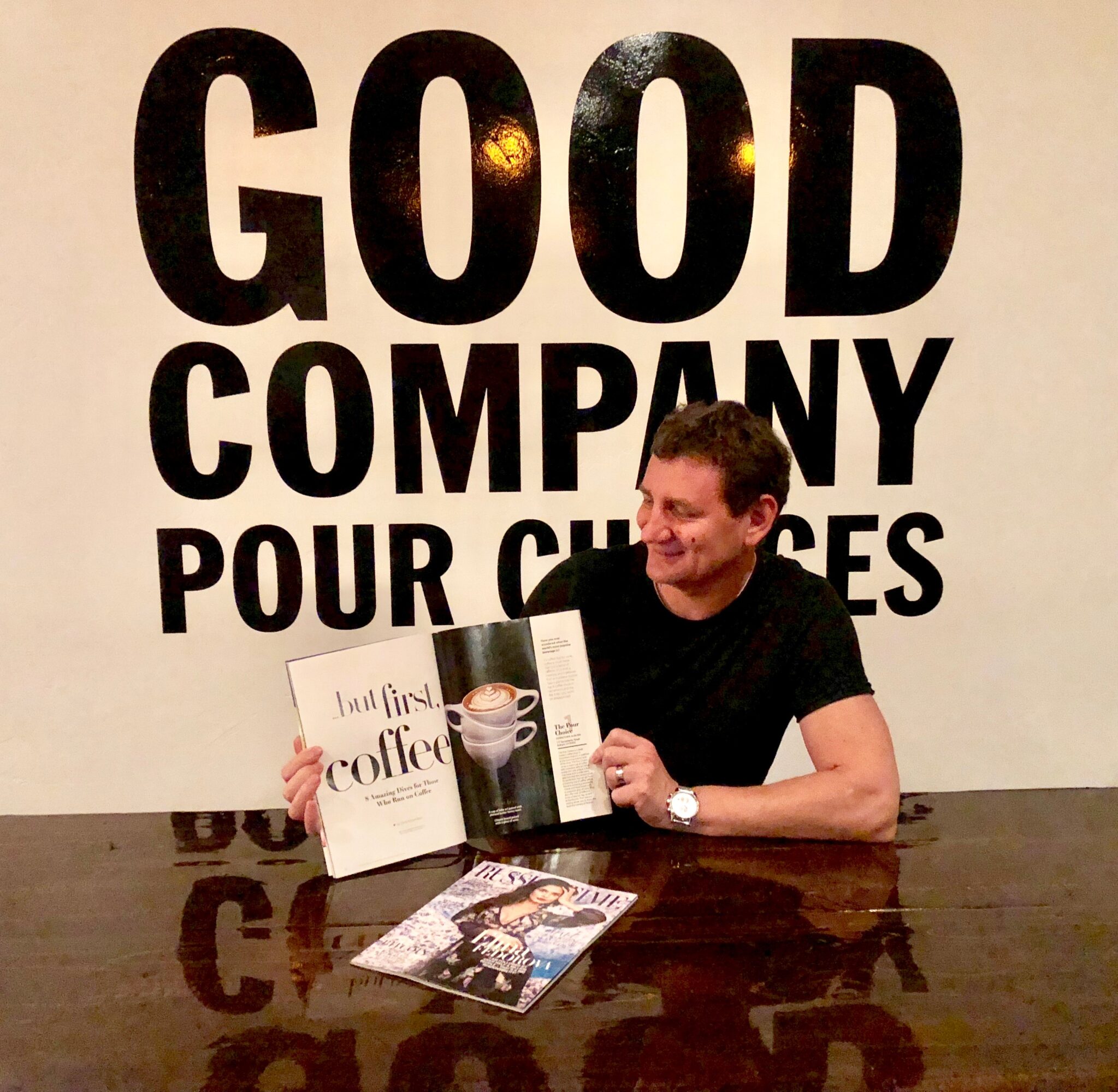 Person sitting at a table reading a magazine, with large text on the wall behind reading 'GOOD COMPANY POUR CHOICES'.