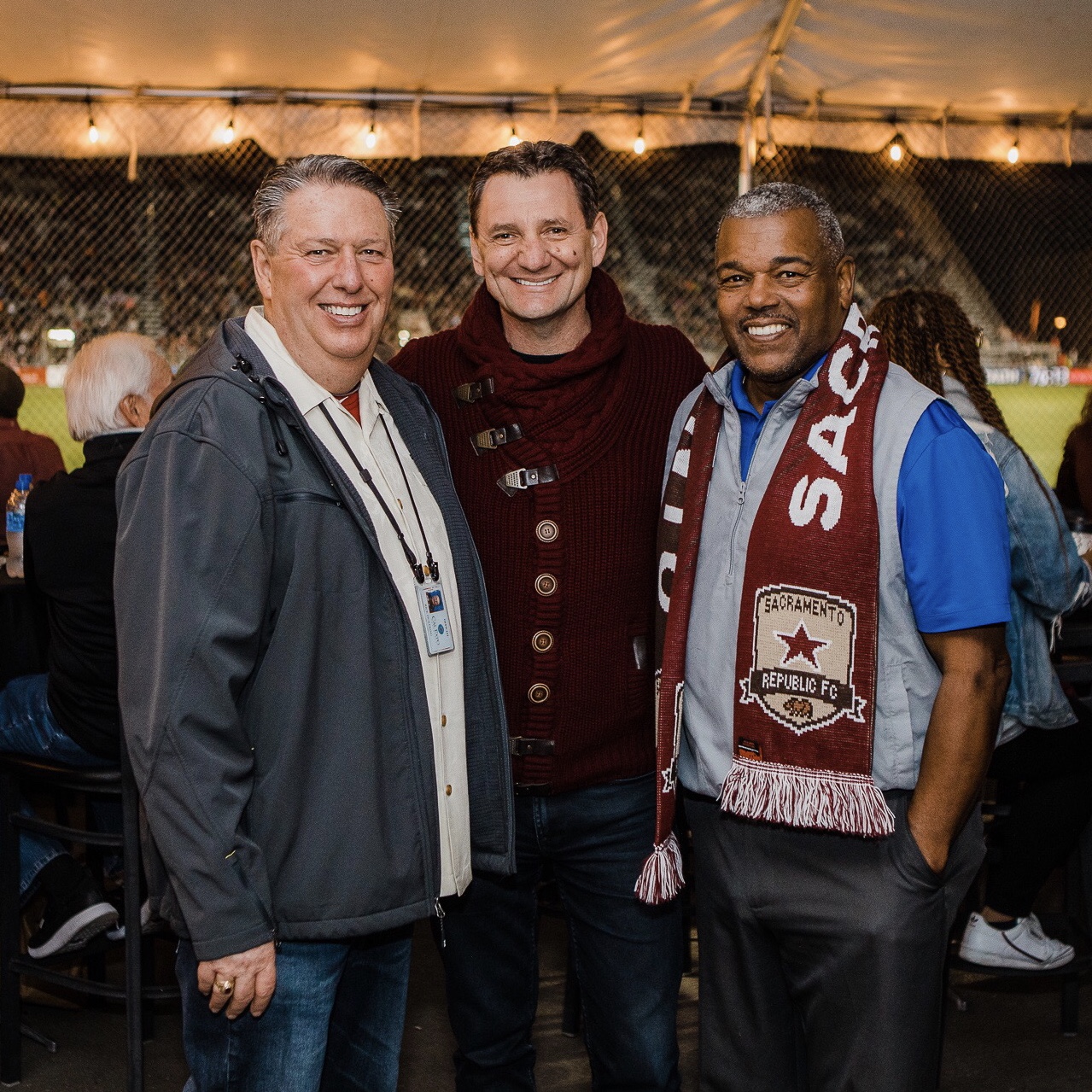 Three men smiling at an outdoor event under a tent, with string lights overhead, standing close together.