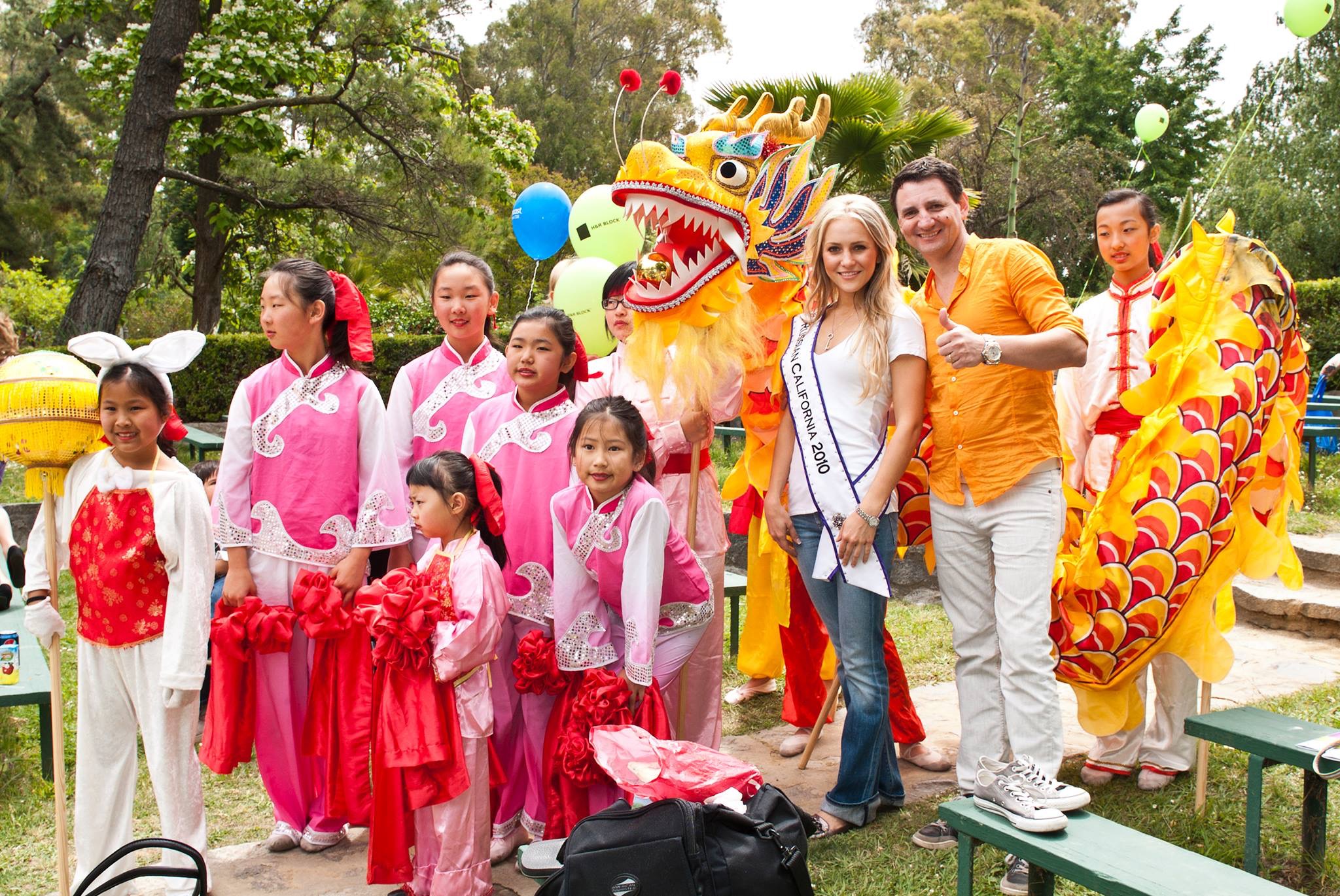 Group of children and adults celebrating outdoors with a colorful dragon costume and balloons, trees in background.