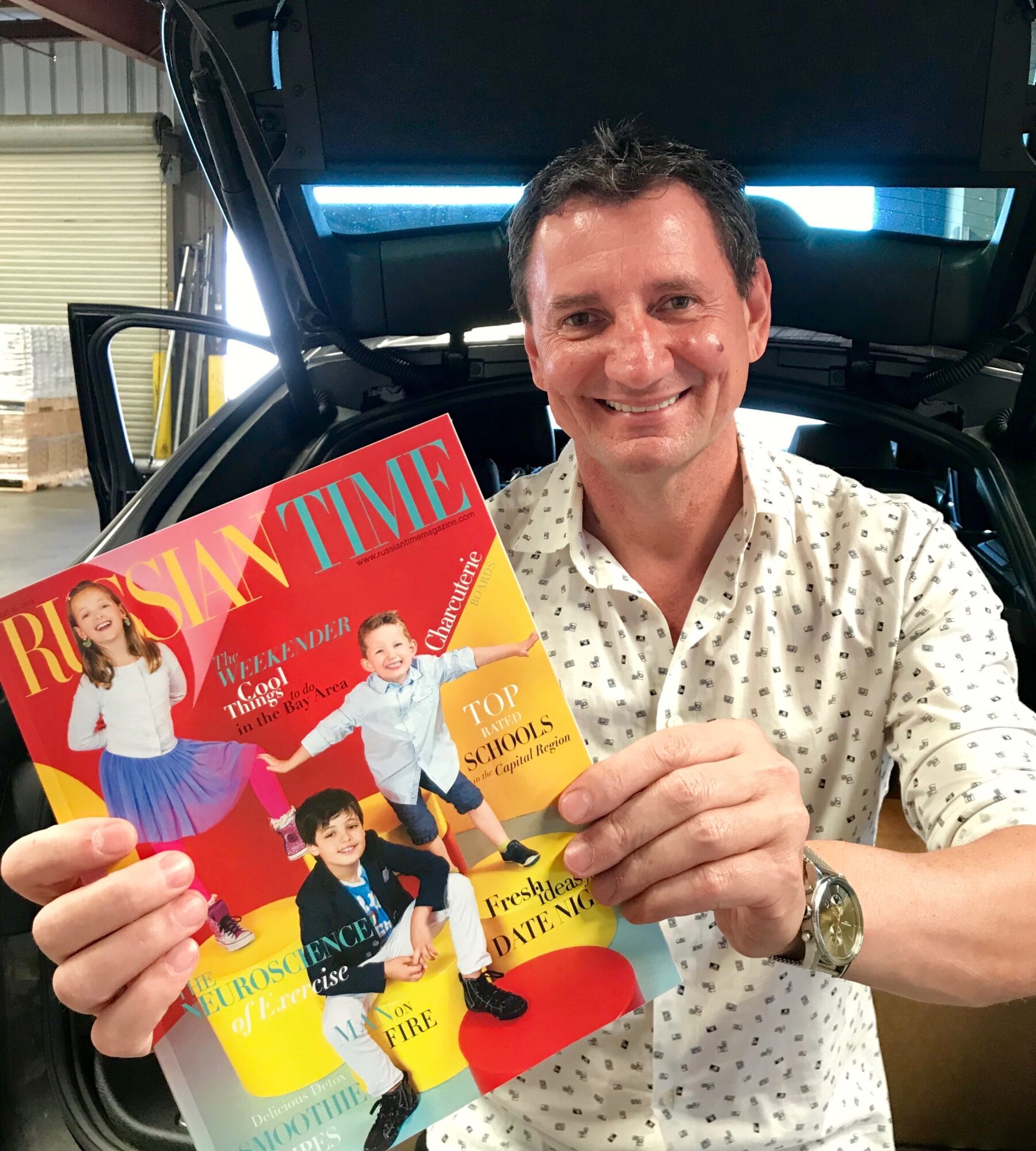 Man smiling holding a magazine in front of a car trunk, with a building in the background.