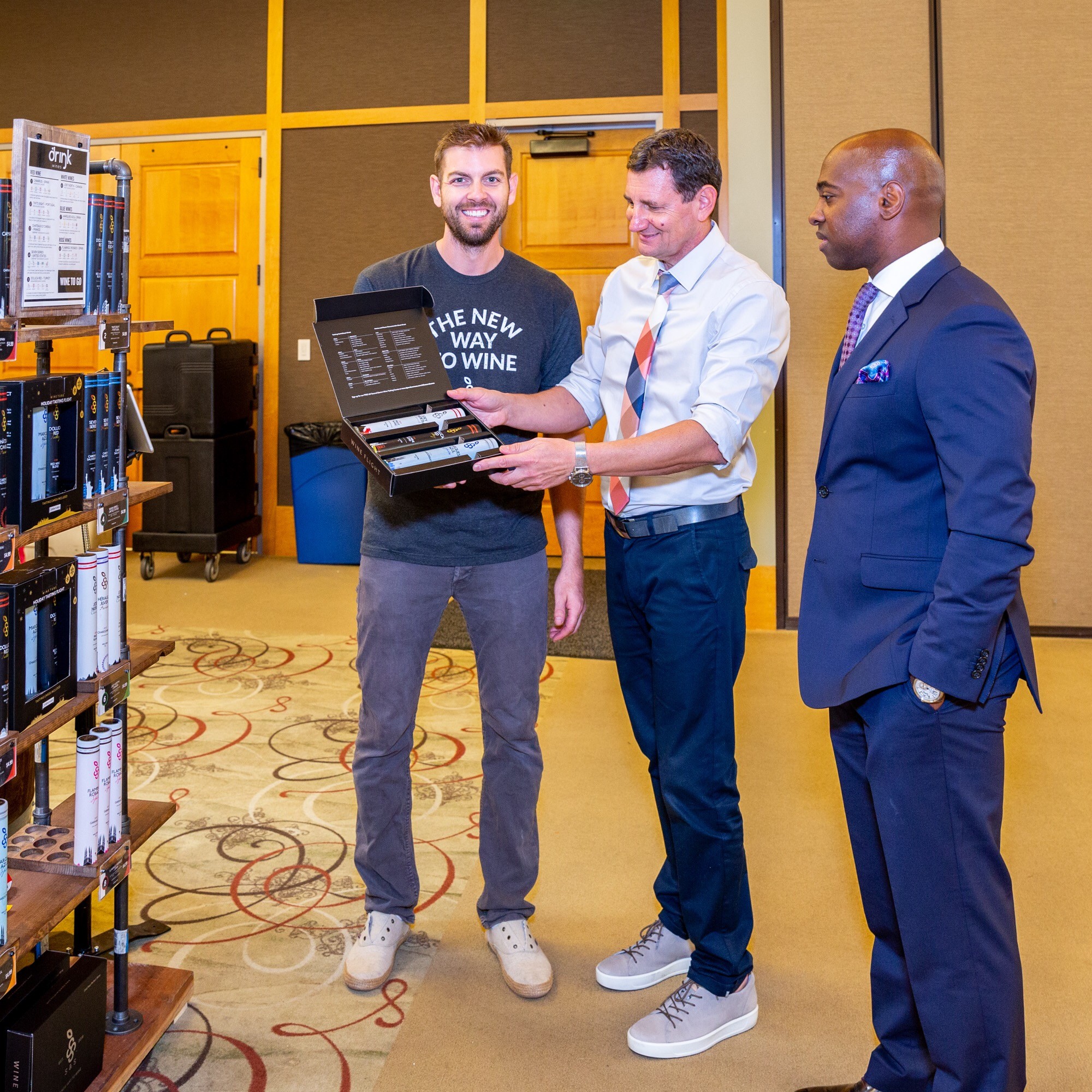 Three men in business attire and casual clothing stand in a room with shelves and a carpeted floor, looking at a laptop. One man shows the laptop to the others, smiling. The room has yellow and brown walls.