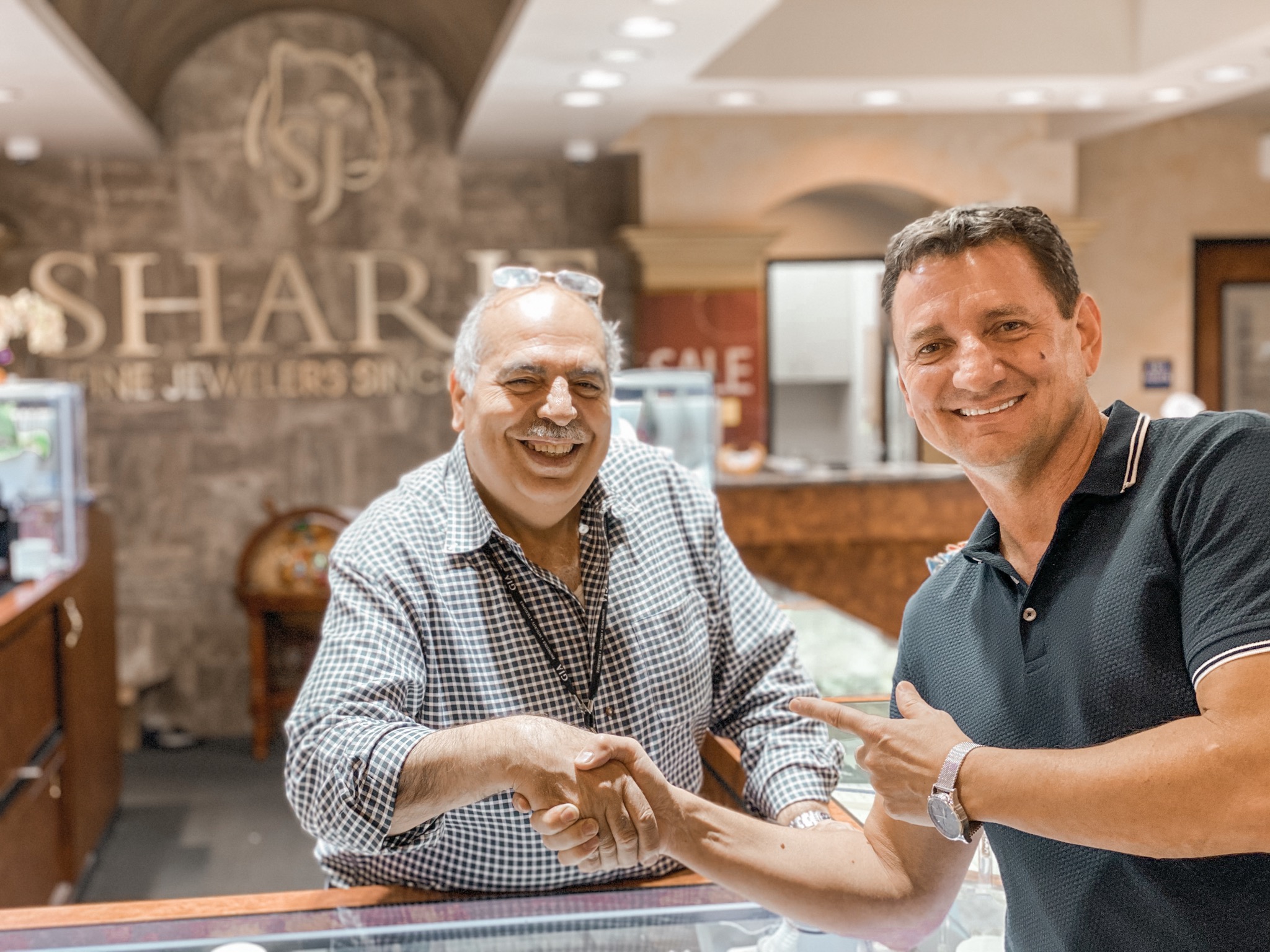Two men shaking hands and smiling inside a jewelry store, with a sign reading 'SHAR' in the background.