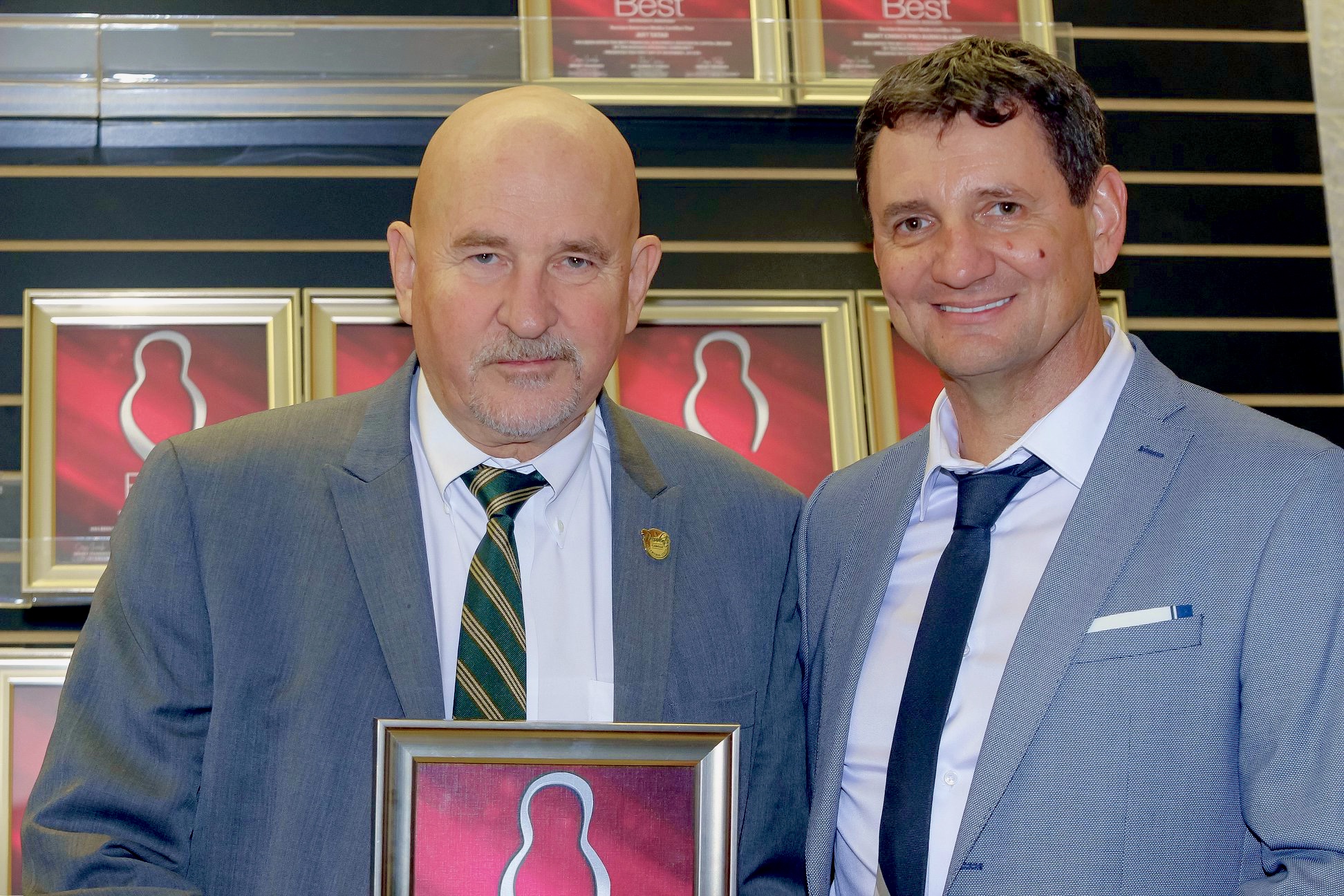 Two men in suits standing indoors, holding a framed picture, smiling, with shelves and framed images in the background.