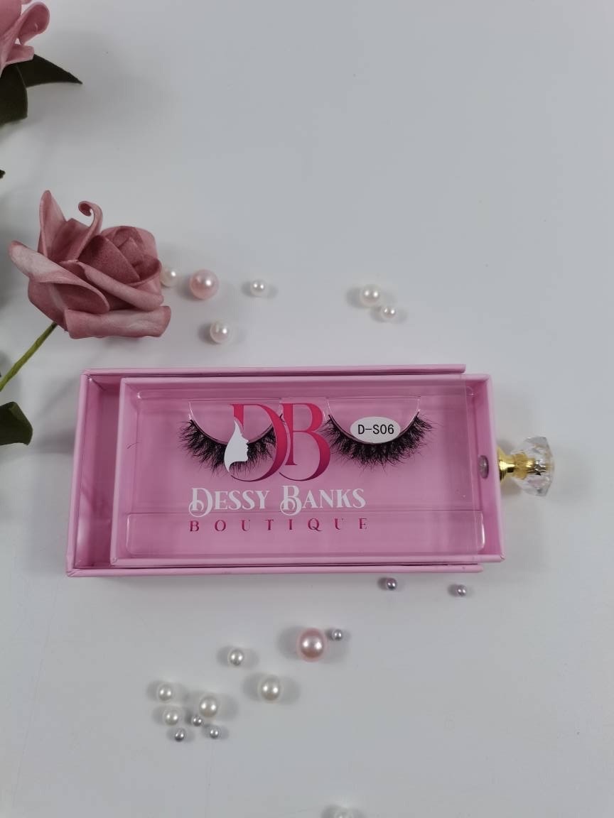Pink rectangular box with a logo and eyelashes, surrounded by pink and white pearls and a pink flower, on a white surface.