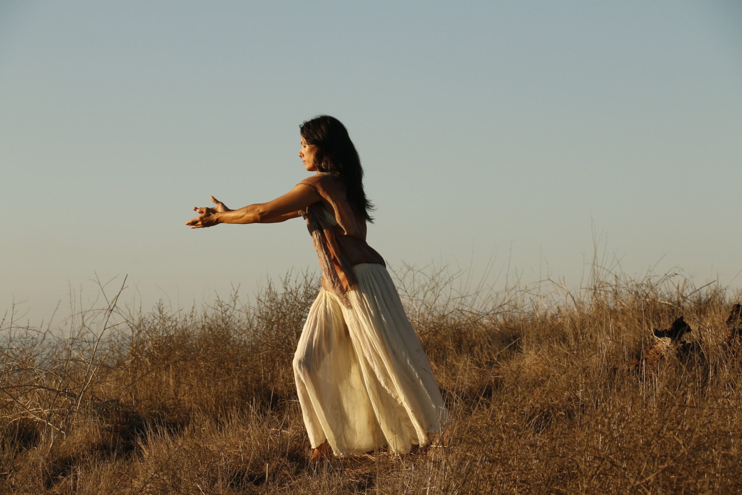 Woman in a long skirt standing in a grassy field with arms extended, facing left, under a clear sky.
