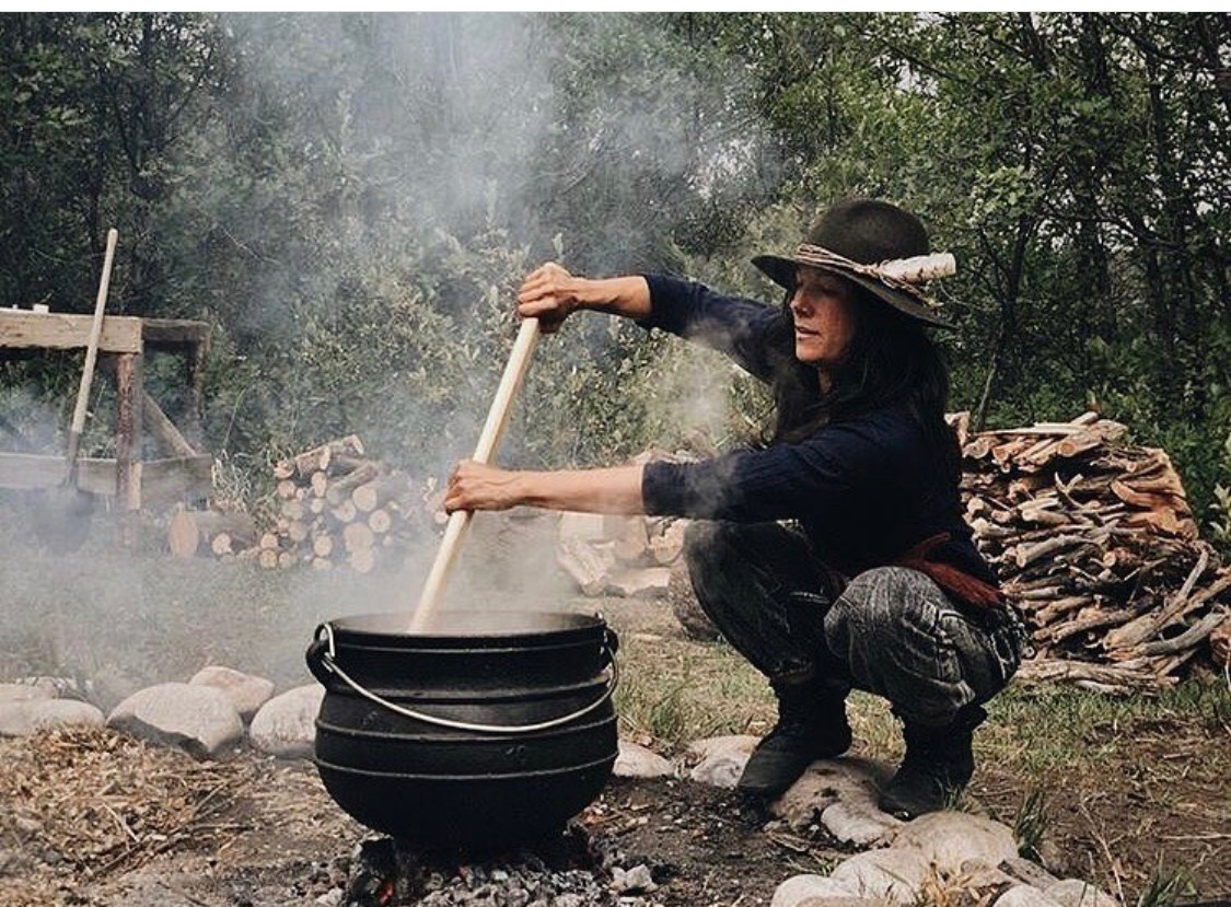 Person crouching outdoors, stirring a large pot over a fire, surrounded by trees and wood logs.