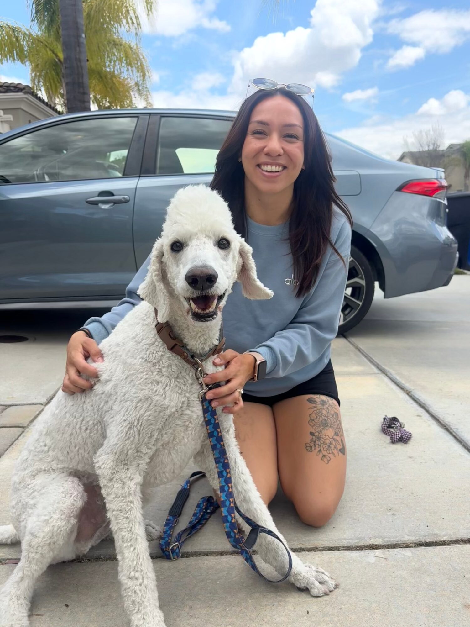 Woman kneeling next to a white dog outdoors, with a car and palm trees in the background, smiling at the camera.