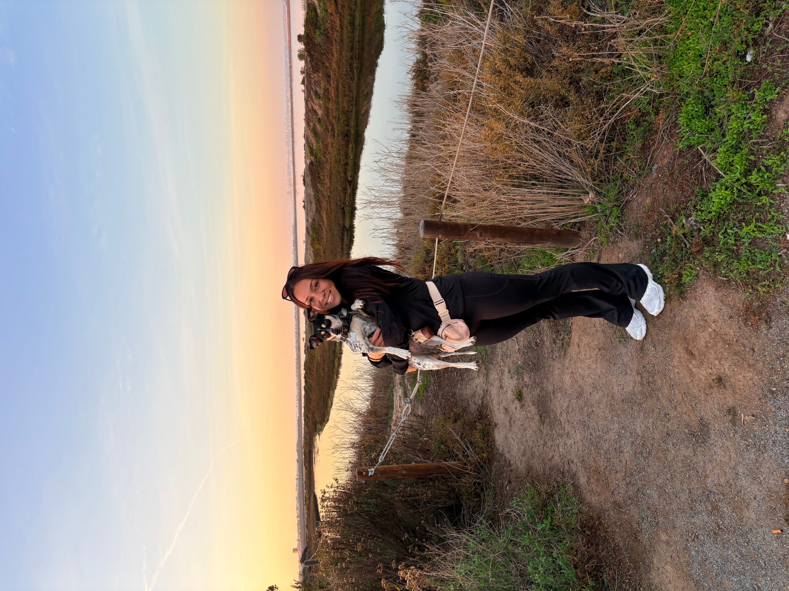 Person standing on a dirt path near a river with a sunset sky, wearing dark clothing and holding a bag.