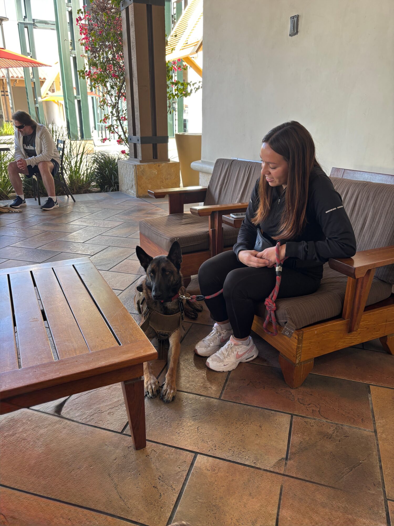 Woman sitting on a wooden bench with a dog on a leash, in a bright indoor space with large windows and plants.
