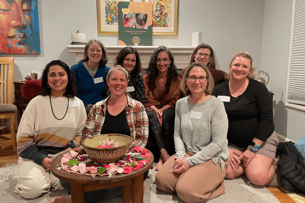 Group of nine women sitting and kneeling around a table with a bowl and pink flowers, smiling indoors.