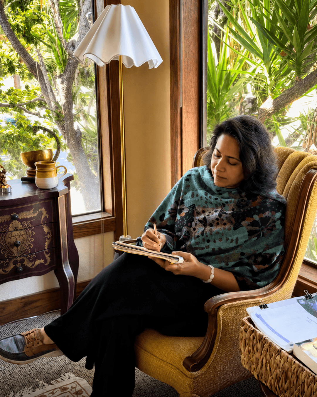 Woman sitting in a yellow armchair, writing in a notebook, near large windows with greenery outside, indoor decor visible.