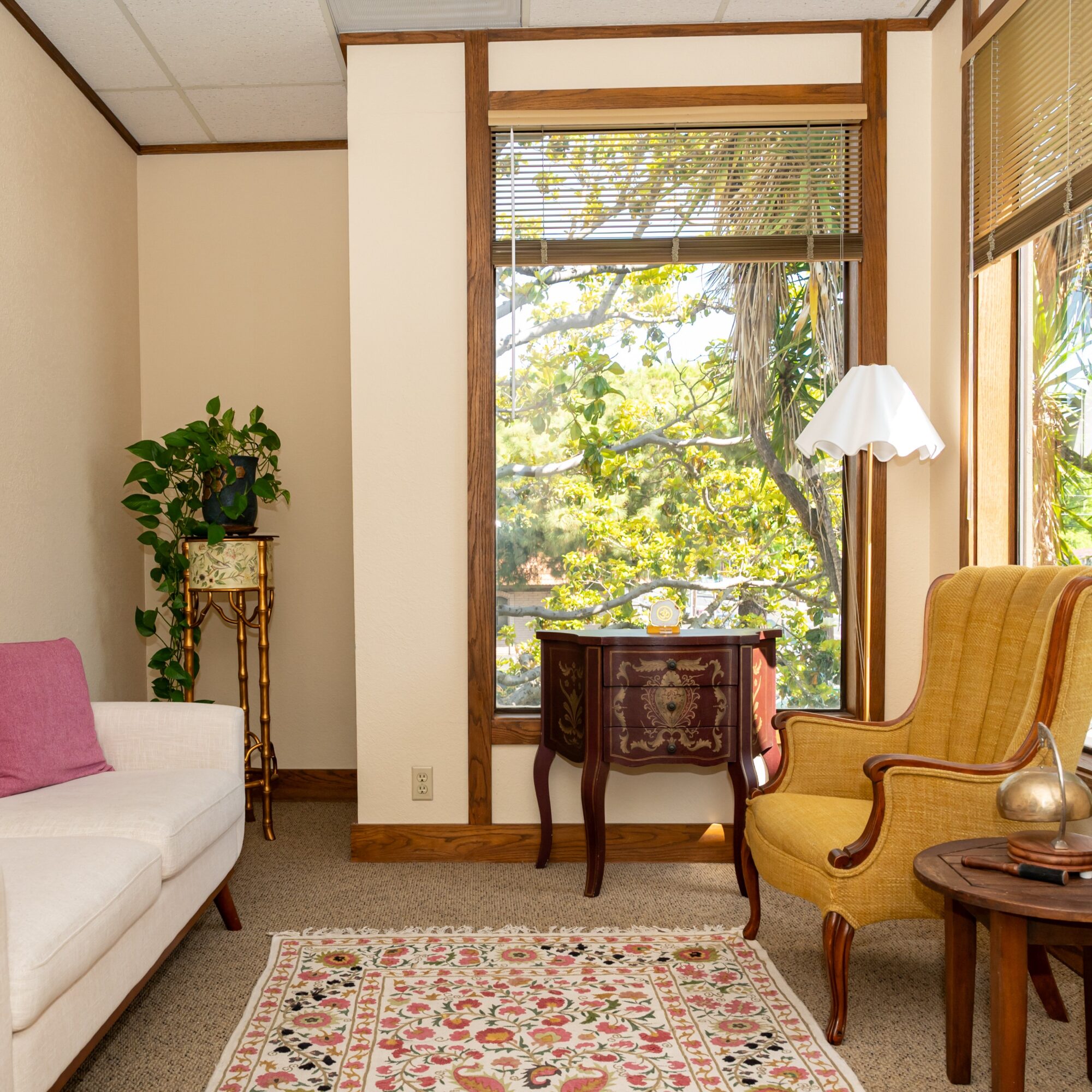 Living room with large window, yellow armchair, white sofa, patterned rug, and potted plant, sunlight streaming through blinds.