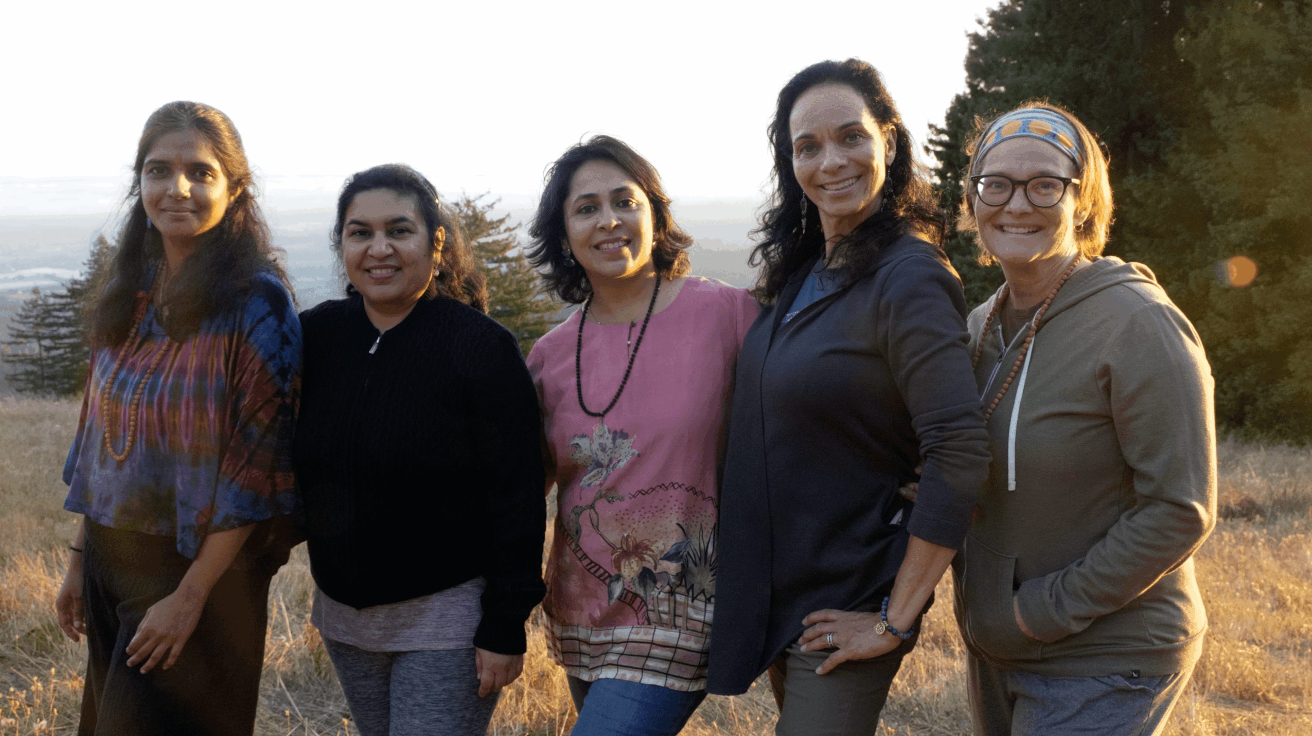 Five women standing outdoors in a line, smiling, with trees and sky in the background.