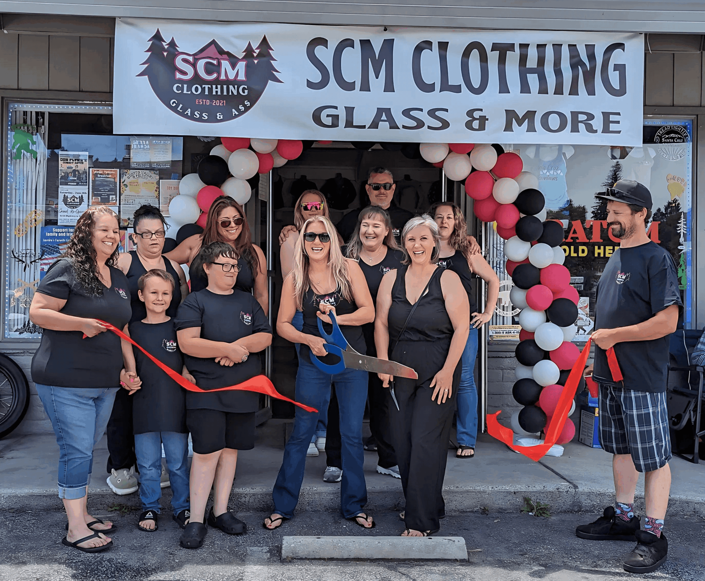 Group of people gathered outside SCM Clothing store for a ribbon-cutting event, with balloons and a banner above the entrance.