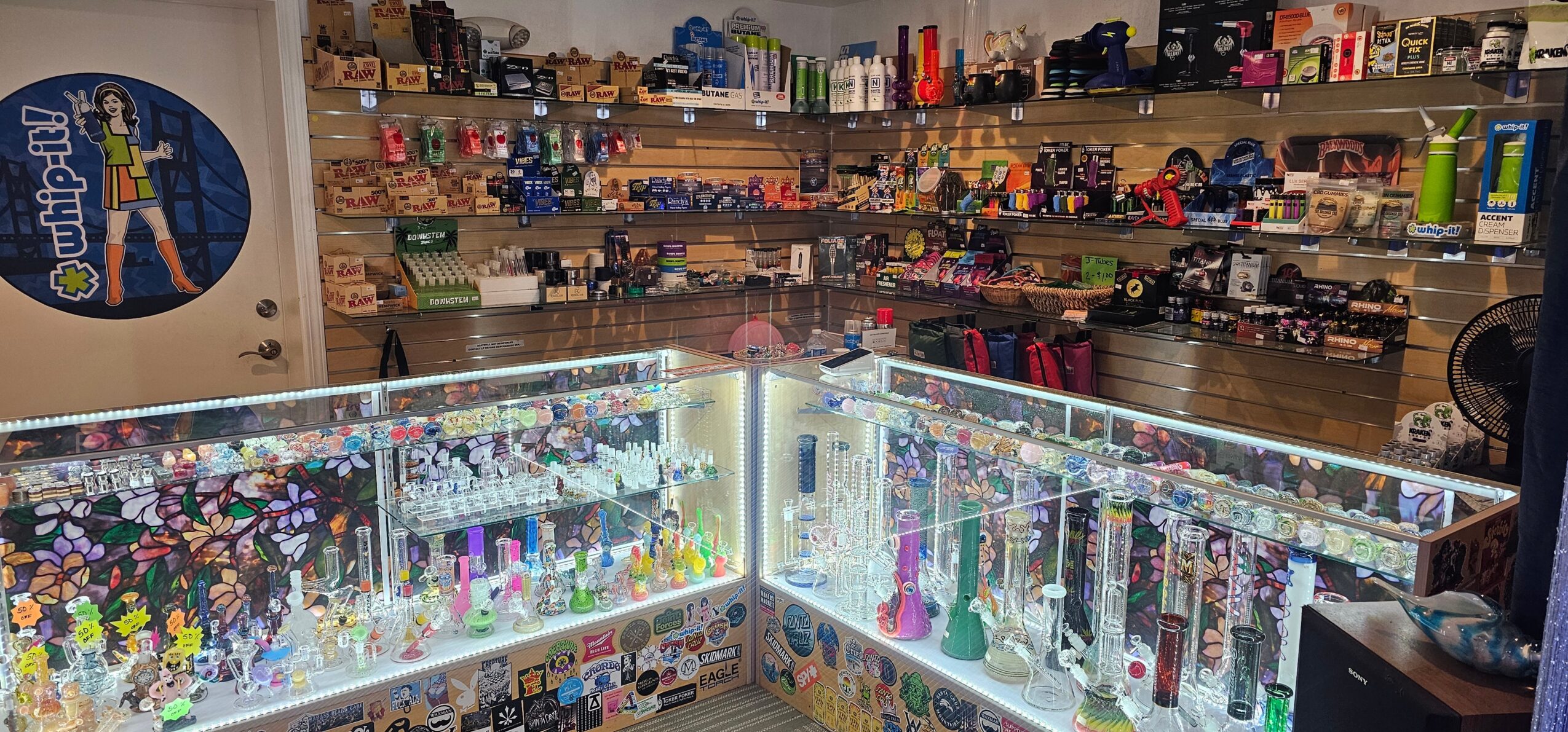 Display case filled with colorful vape pens and accessories in a store, shelves with various products in background.