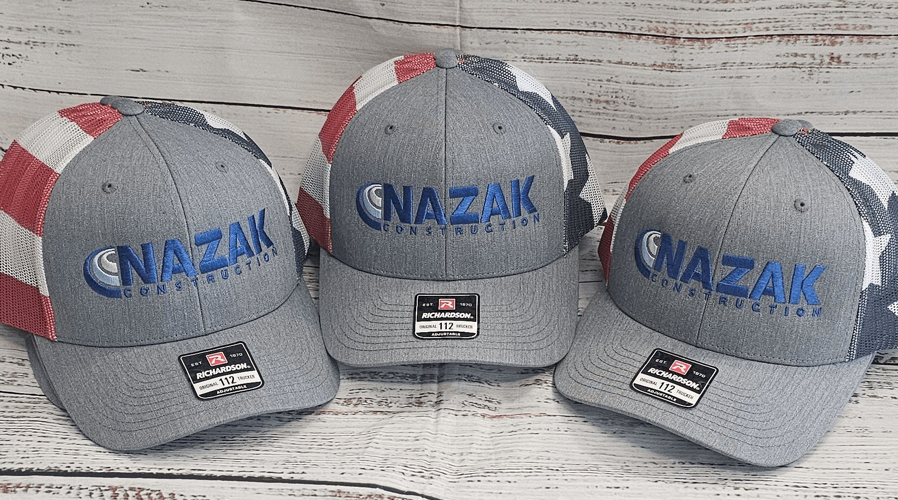 Three gray baseball caps with blue and red accents and the word 'NAZAK' embroidered on the front, placed on a wooden surface.