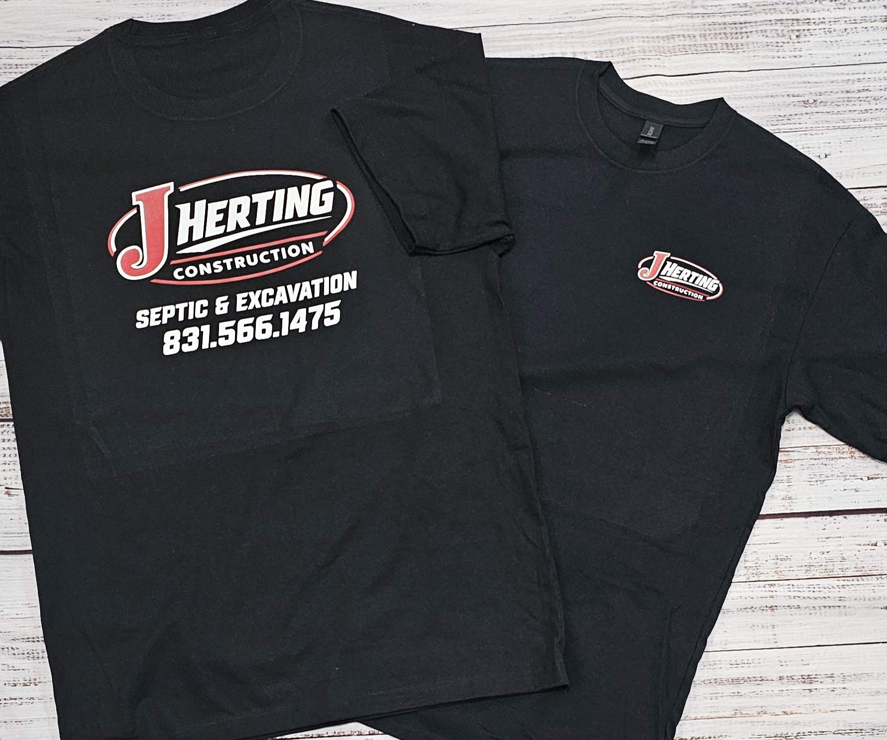 Two black T-shirts with company logos and contact information, laid flat on a white wooden surface.