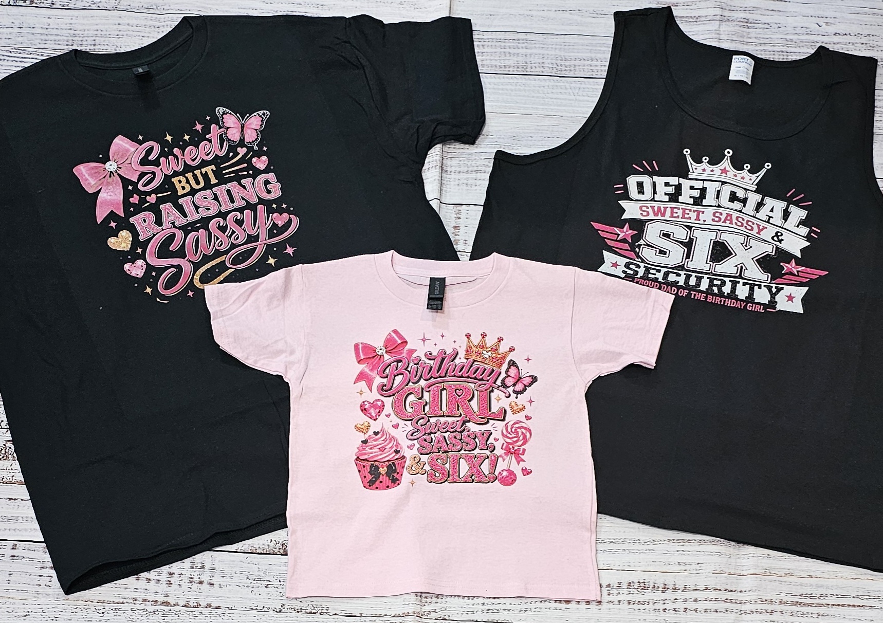 Three children's T-shirts with pink, black, and white colors, featuring playful text and graphics, laid on a wooden surface.