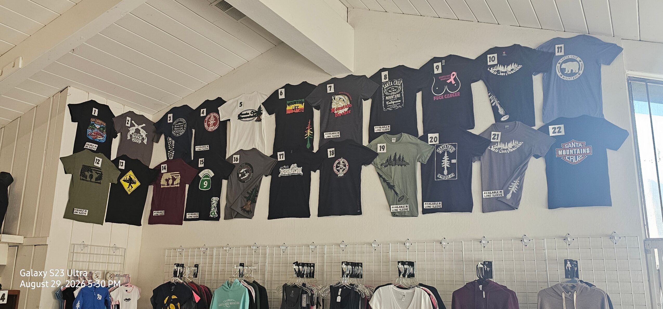 T-shirts displayed on a wall, arranged in rows, with some hanging on hooks below. The wall is white with a sloped ceiling.