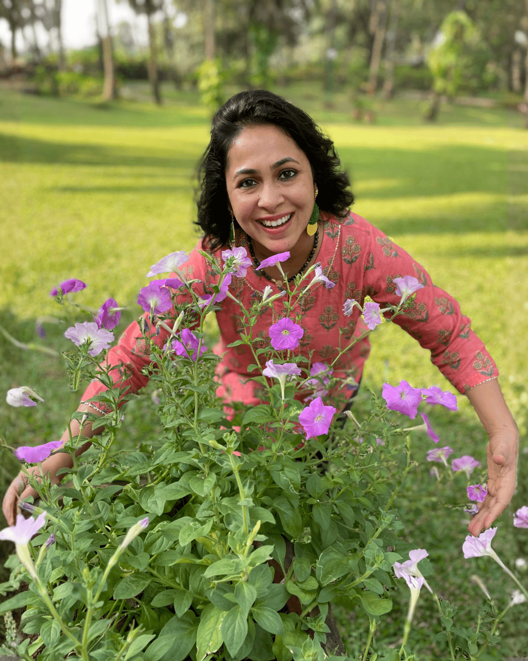 Smiling woman in pink dress crouching behind purple and white flowers in a park.