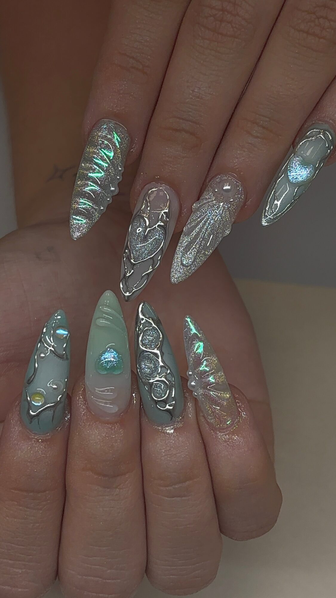 Close-up of ten decorated fingernails with holographic, metallic, and 3D embellishments, featuring various designs and textures.