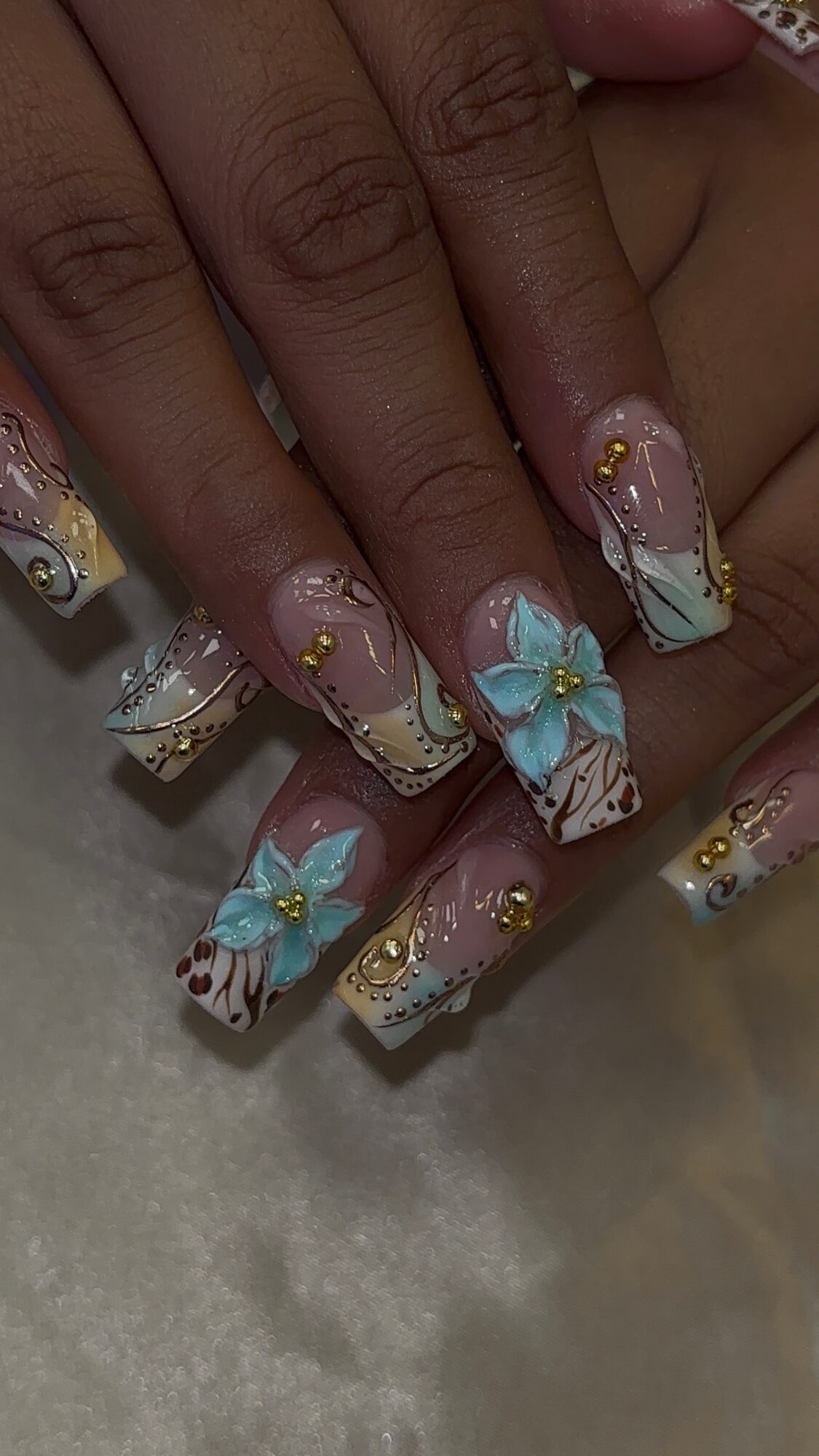 Nail art with floral design, gold accents, and a large blue flower on a light pink background.