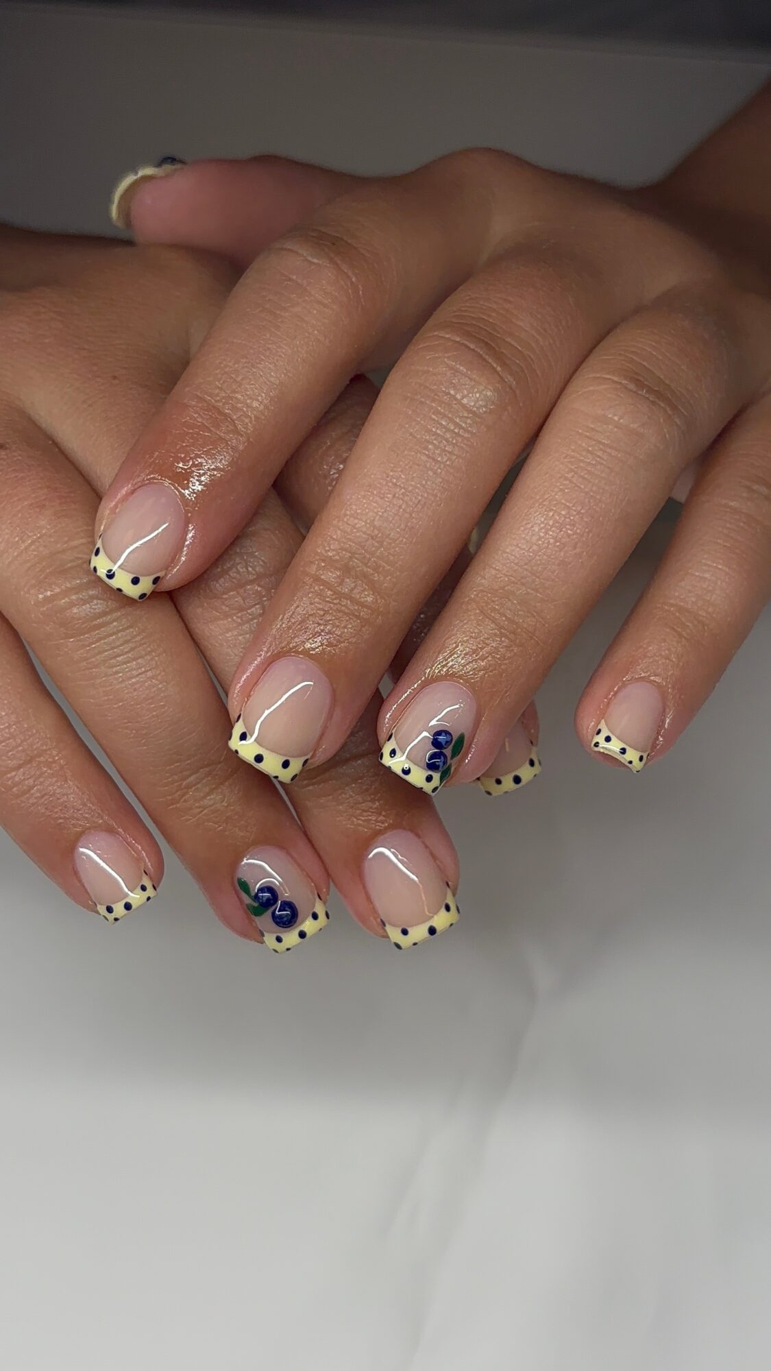 Hands with decorated nails featuring black, blue, and white dots and patterns, resting on a white surface.