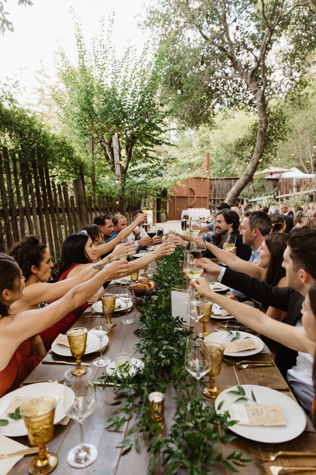People sitting at a long outdoor table raising glasses in a toast, surrounded by trees and greenery.