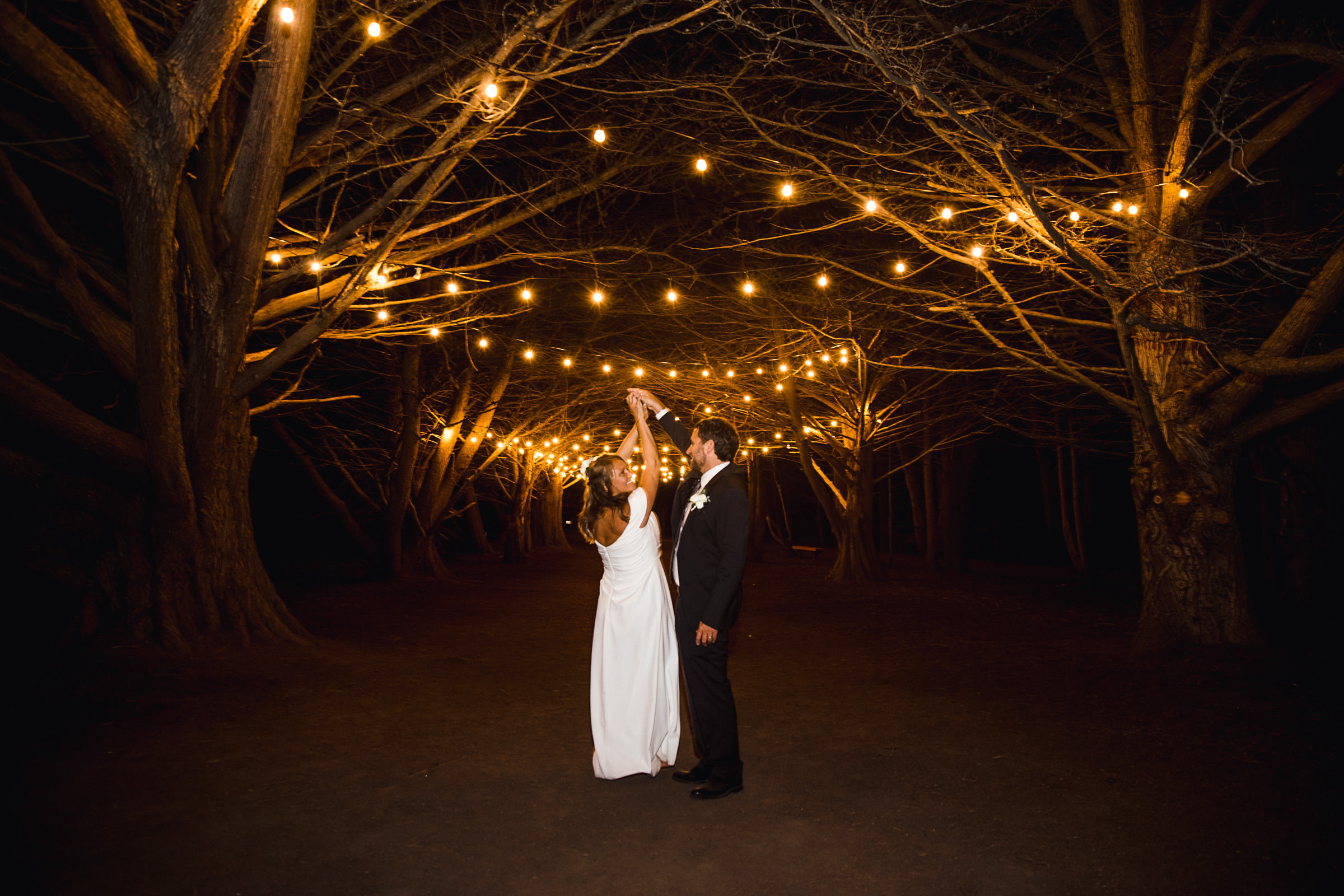 A couple stands under string lights hanging between trees at night, holding hands and looking at each other.
